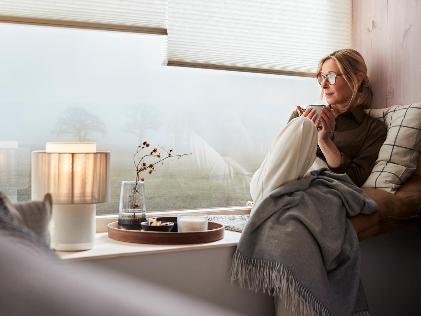 A woman taking a break at home sitting in a window seat. She’s relaxing with tea, SYMFONISK speaker lamp and HOLMVI throw.