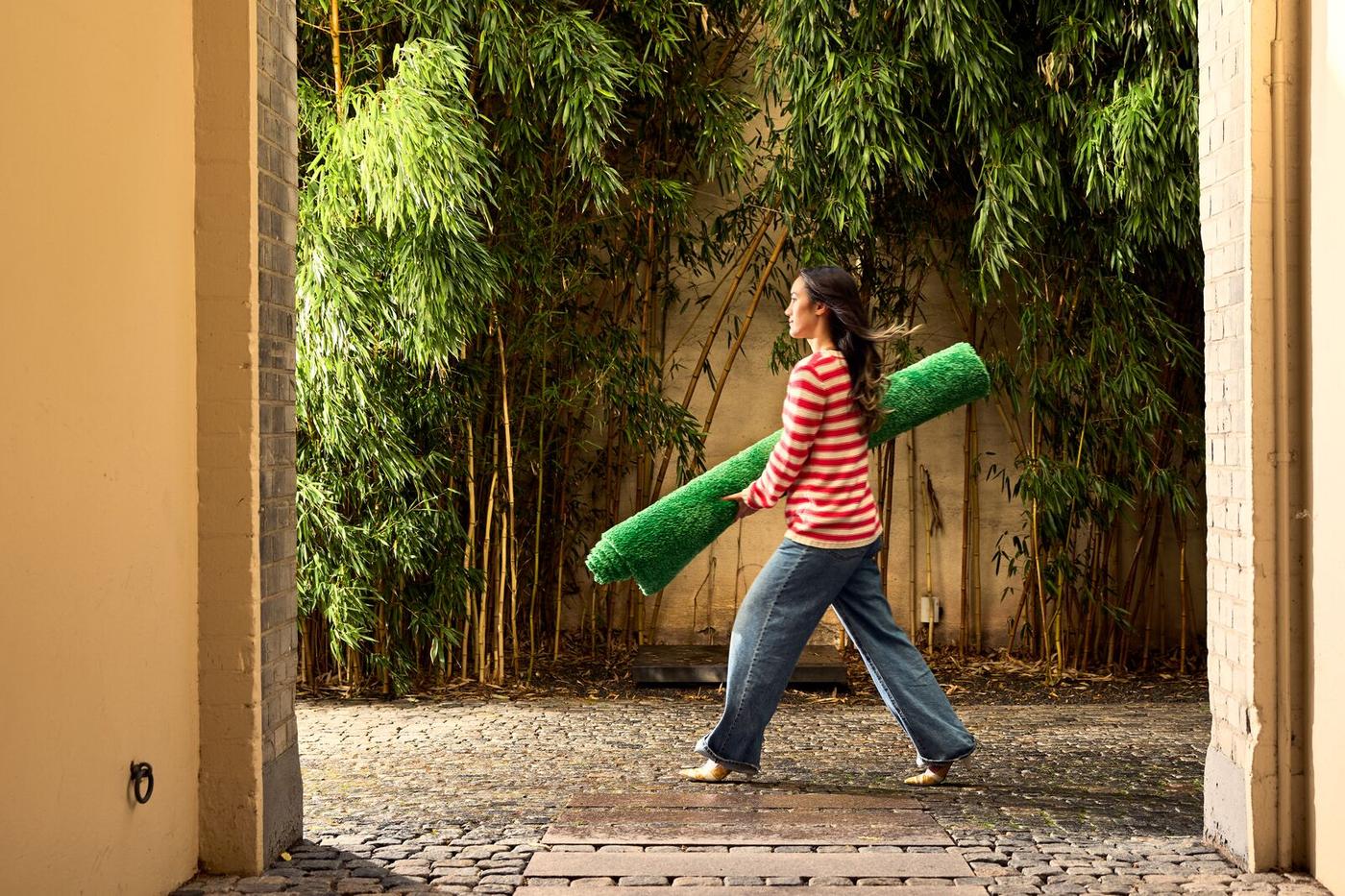 A woman walking through a courtyard with a stone floor, surrounded by light-colored walls and bamboo plants. She is carrying a roll of green artificial grass while wearing a red and white striped T-shirt and jeans.
