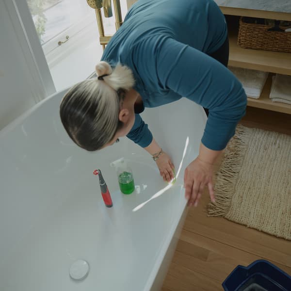 A woman wearing a blue long sleeve t-shirt is cleaning a white bathtub. 