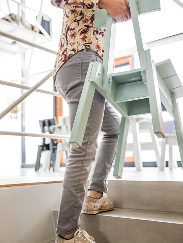 A woman wearing a floral print shirt, jeans and sneakers, is carrying a mint-coloured wooden chair up the stairs.