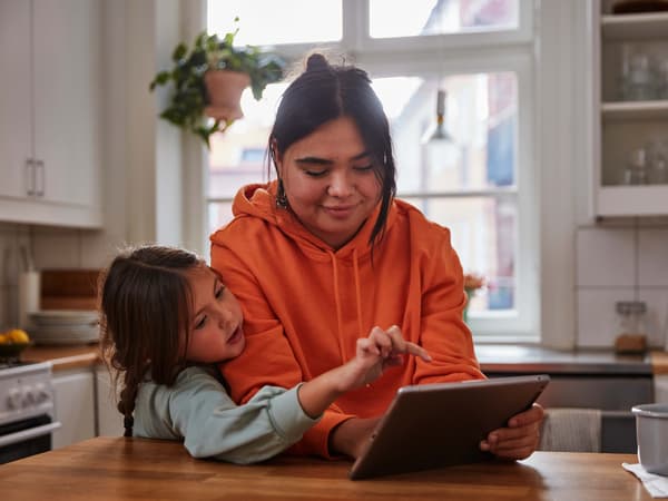 A woman wearing an orange jumper stands in a kitchen next to a little girl, they are using a tablet.