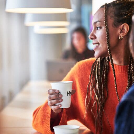 A woman wearing an orange sweater is holding a cup while looking out the window.
