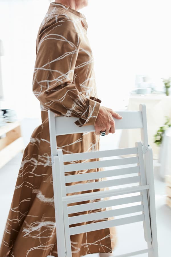 A woman with a brown patterned dress carrying a TERJE chair in white wood in a white setting.