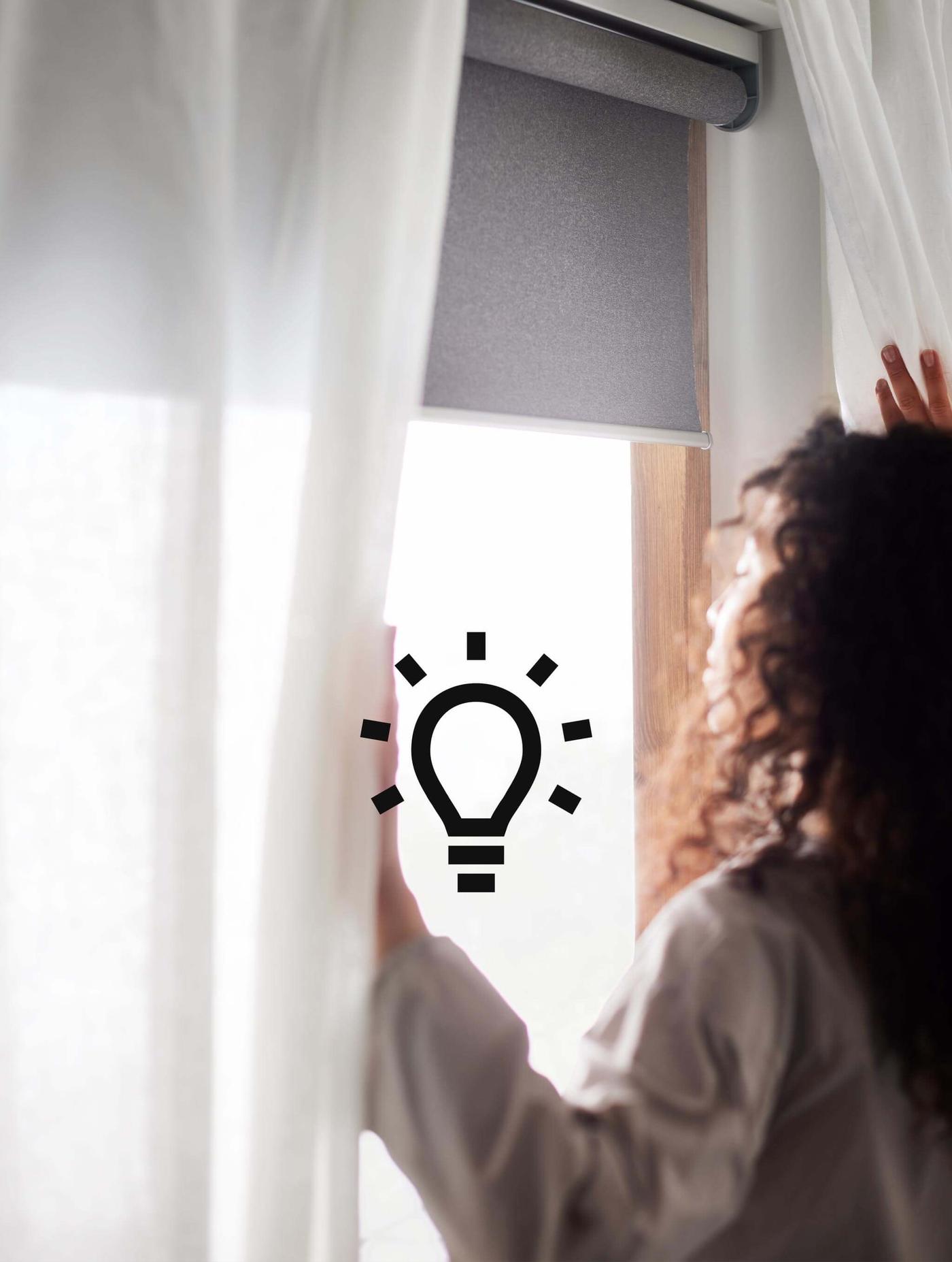 A woman with brown curly hair opening blinds and a blackout roller blind with the sun shining in and a light bulb icon on the image.