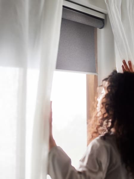 A woman with brown curly hair opening blinds and a blackout roller blind with the sun shining in.