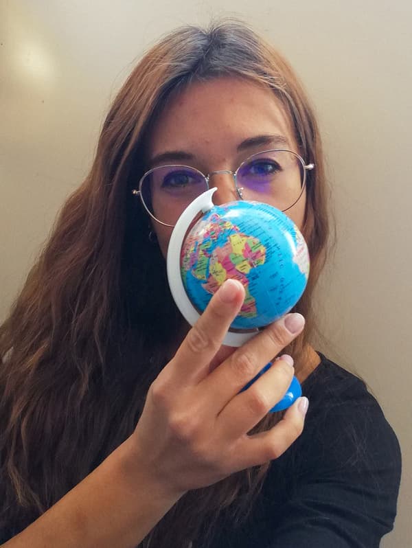 A woman with brown hair, wearing glasses is holding a small, plastic globe in front of her face.