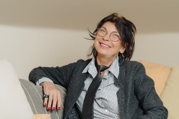 A woman with dark, shoulder-length hair and glasses, dressed in a classic-style suit and tie, is sitting on the sofa, leaning back against the cushions and smiling.