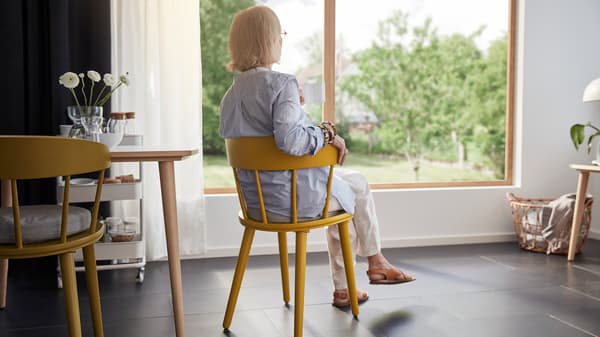 A woman with grey hair sits in a yellow chair in a room looking out through a big window.