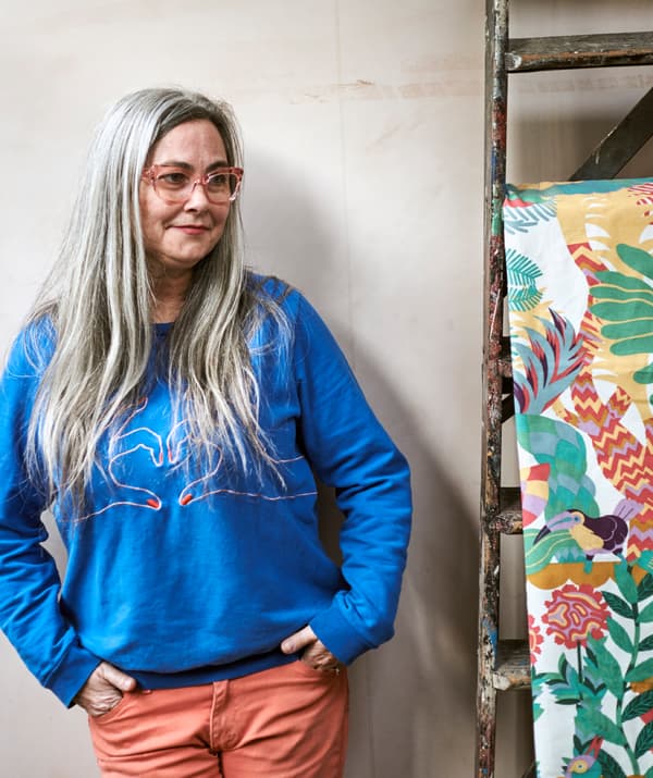 A woman with long grey hair in pink-framed glasses and blue sweatshirt next to brightly patterned fabric hung on a ladder.