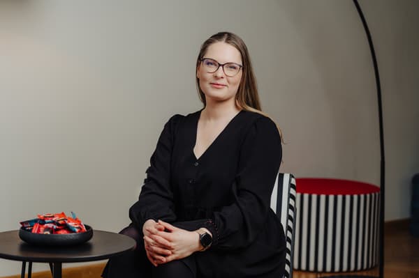 A woman with long hair and glasses, wearing a black dress, is sitting in a chair and looking at the camera.