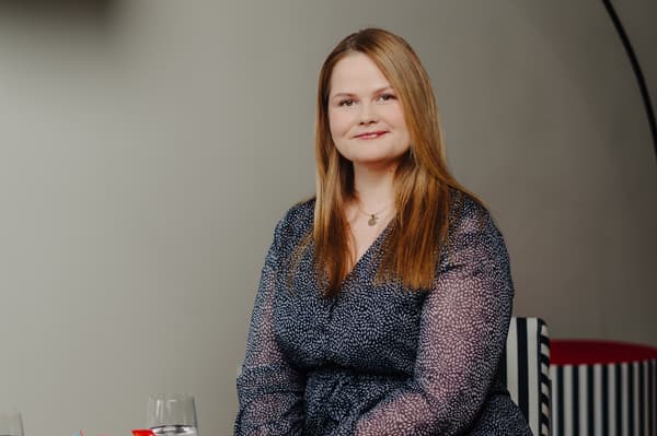 A woman with longish, light brown hair, wearing a black-and-white dress, is sitting in a chair and looking at the camera.