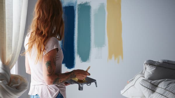 A woman with red hair is standing in a bedroom trying out different paint colours by painting lines on a white wall.