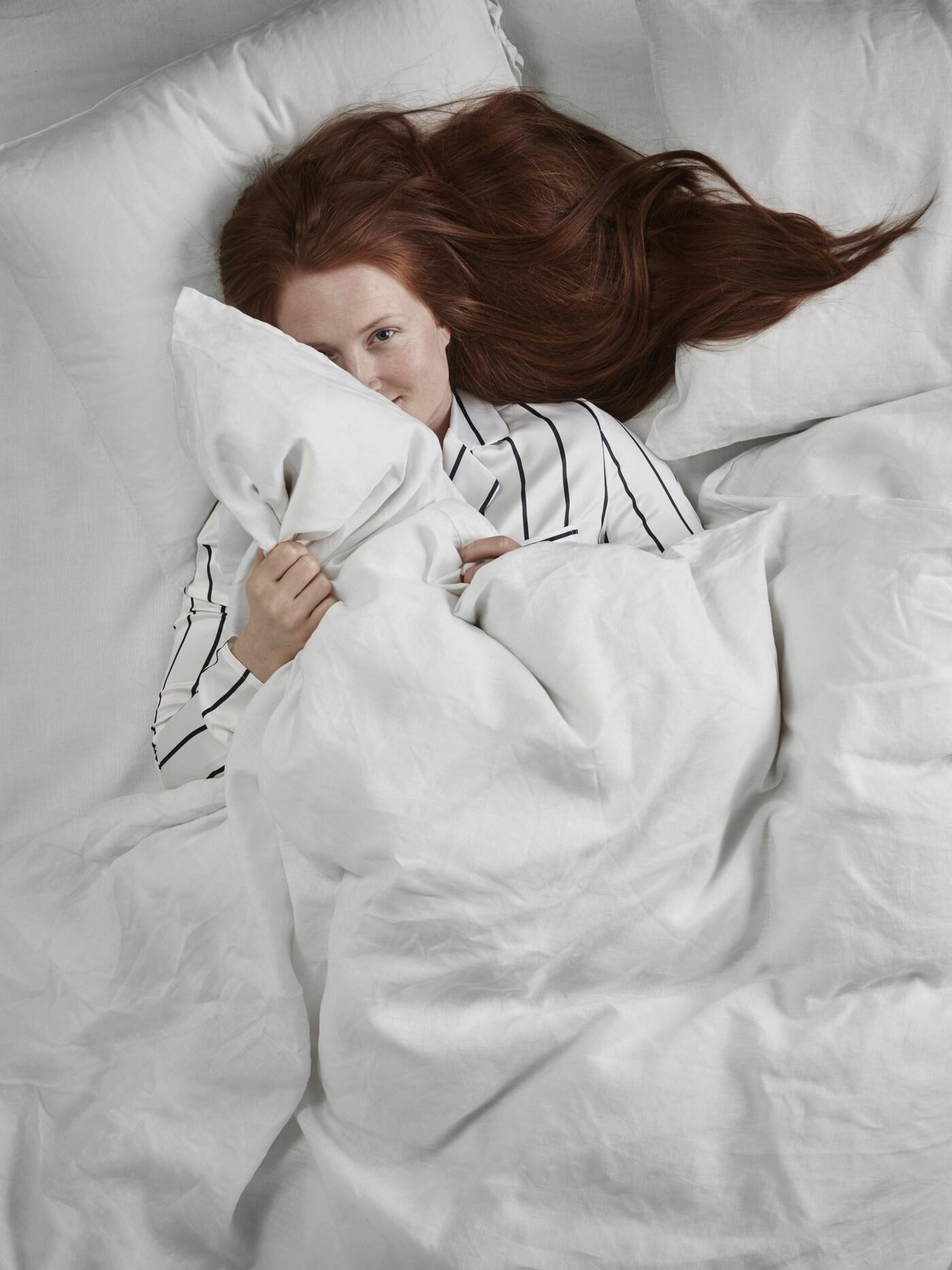 A woman with red hair laying in a bed with a white comforter and white pillows.
