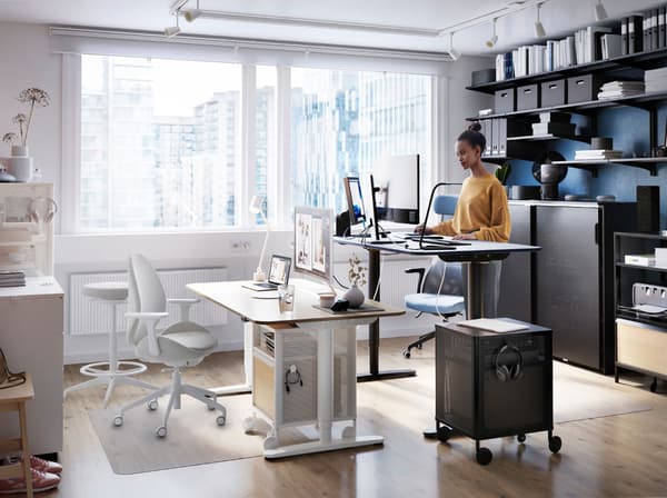 A woman working at a BEKANT sit/stand desk, beside a large window. ALGOT shelves holding files are on the wall behind her.