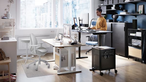 A woman working at a BEKANT sit/stand desk, beside a large window, shelves holding files are on the wall behind her.