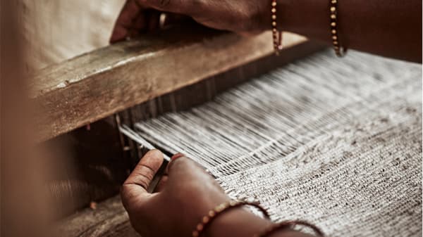 A woman’s is busy weaving a piece of beige textile with her hands. She is wearing golden bangles around her arms.