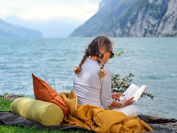 A women reading a book, while sitting by the lake on a mustard colored blanket 