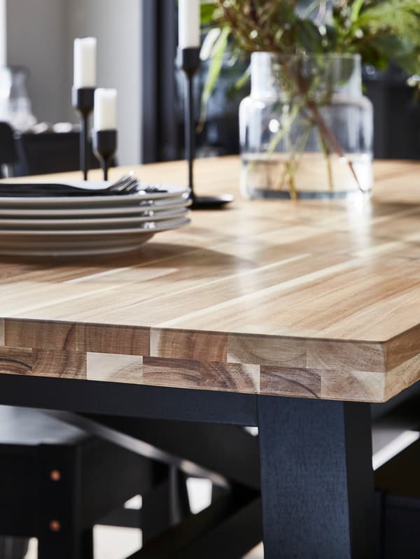 A wood-topped dining table with a striking grain pattern upon which is a pile of dinner plates, some candlesticks and a vase.