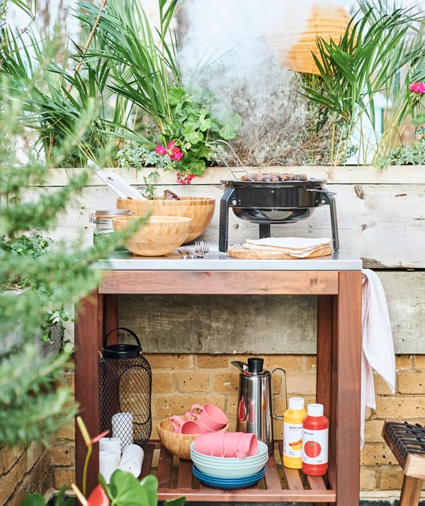 A wood trolley in an outdoor cooking area with a BBQ on its metal worktop and eatware and condiments on a shelf below.