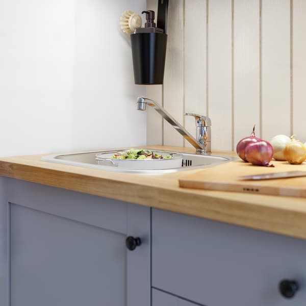 A wood worktop, grey kitchen fronts, a stainless steel inset sink, a chrome-plated kitchen mixer tap and salad in a colander.