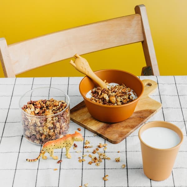A wooden board with a ceramic bowl of breakfast cereal and milk on the table. Next to it - a glass of milk, a container of breakfast cereal, and a handful of cereal scattered on the table which a tiger figurine seems to be enjoying.