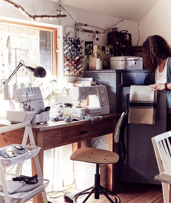 A wooden desk and swivel chair in front of a large window with sewing machines and craft supplies.
