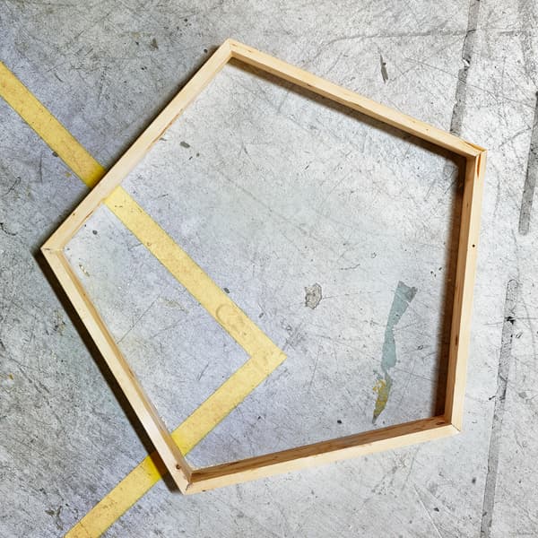 A wooden frame in the shape of a pentagon lying on a scuffed concrete floor.