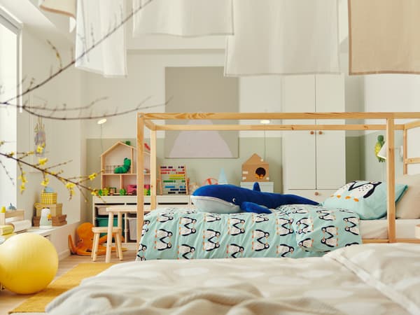 A wooden KURA bed is in the middle of a large children’s bedroom. A giant stuffed whale sits on top of the duvet.