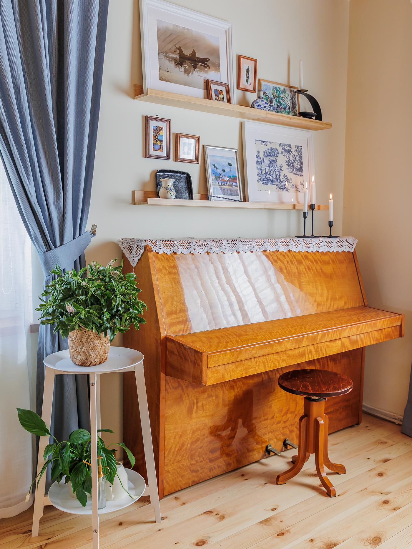 A wooden piano with a cushioned bench, topped by two white wall shelves adorned with framed pictures and small decorative items.
