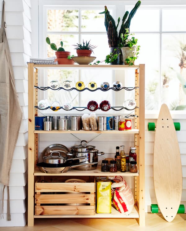 A wooden shelving unit with two bottle racks. Cookware, cans, jars, bottles and green plants are stored/standing here.