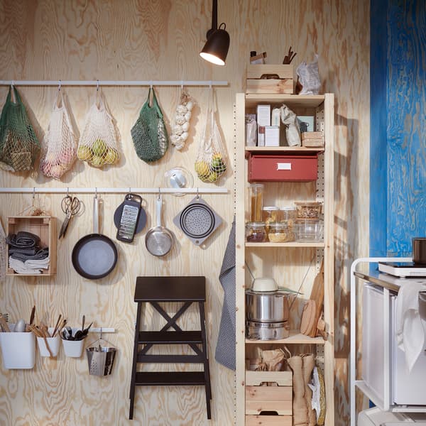 A wooden wall where white rails with hooks are mounted: net bags with fruits, kitchen utensils and a step stool hang here.