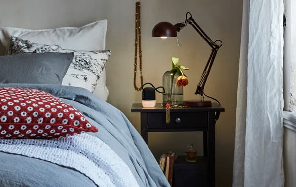 A work lamp on a table next to a bed shines light on a glass vase and alarm clock in a cosy bedroom.