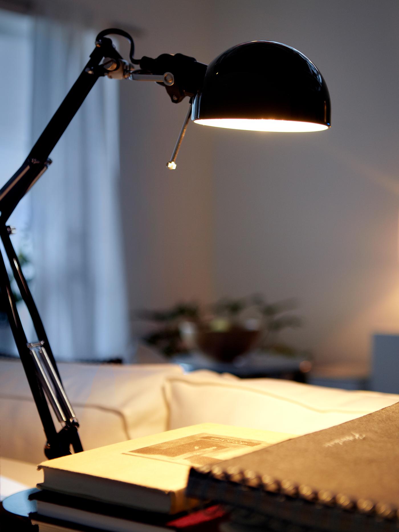 A work lamp shining down onto some books on a table, with a sofa and a window with curtains in the background.