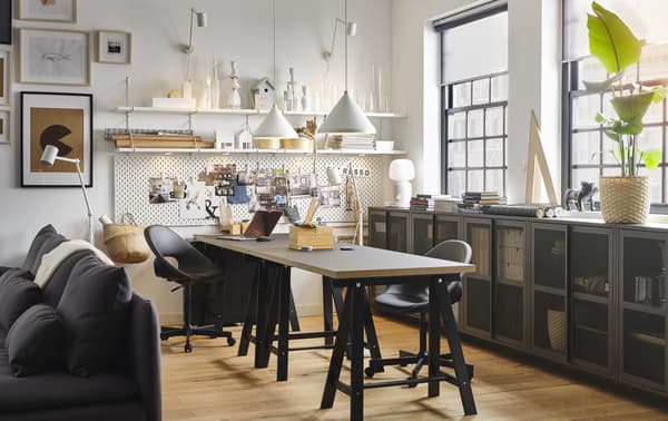 A workspace with two black tables and two black swivel chairs, two white pendant lamps and white wall shelves.