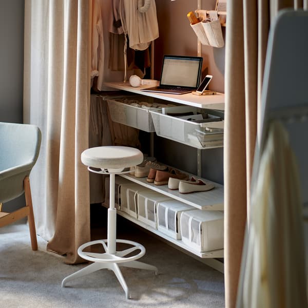 A workstation integrated into a white BOAXEL storage system behind curtains, with lighting and a sit/stand support.