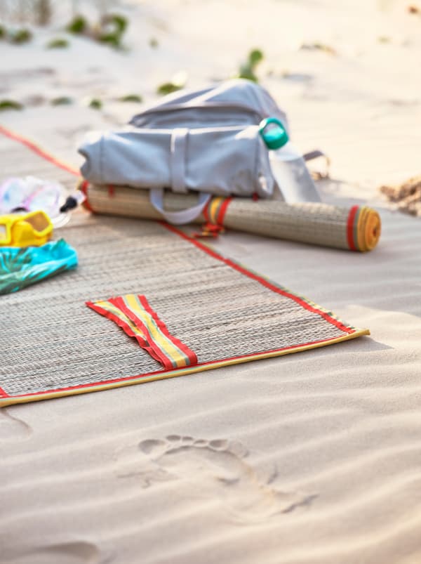 A woven beach mat with yellow and orange trimmings lying on sand. A light grey backpack lies next to it.