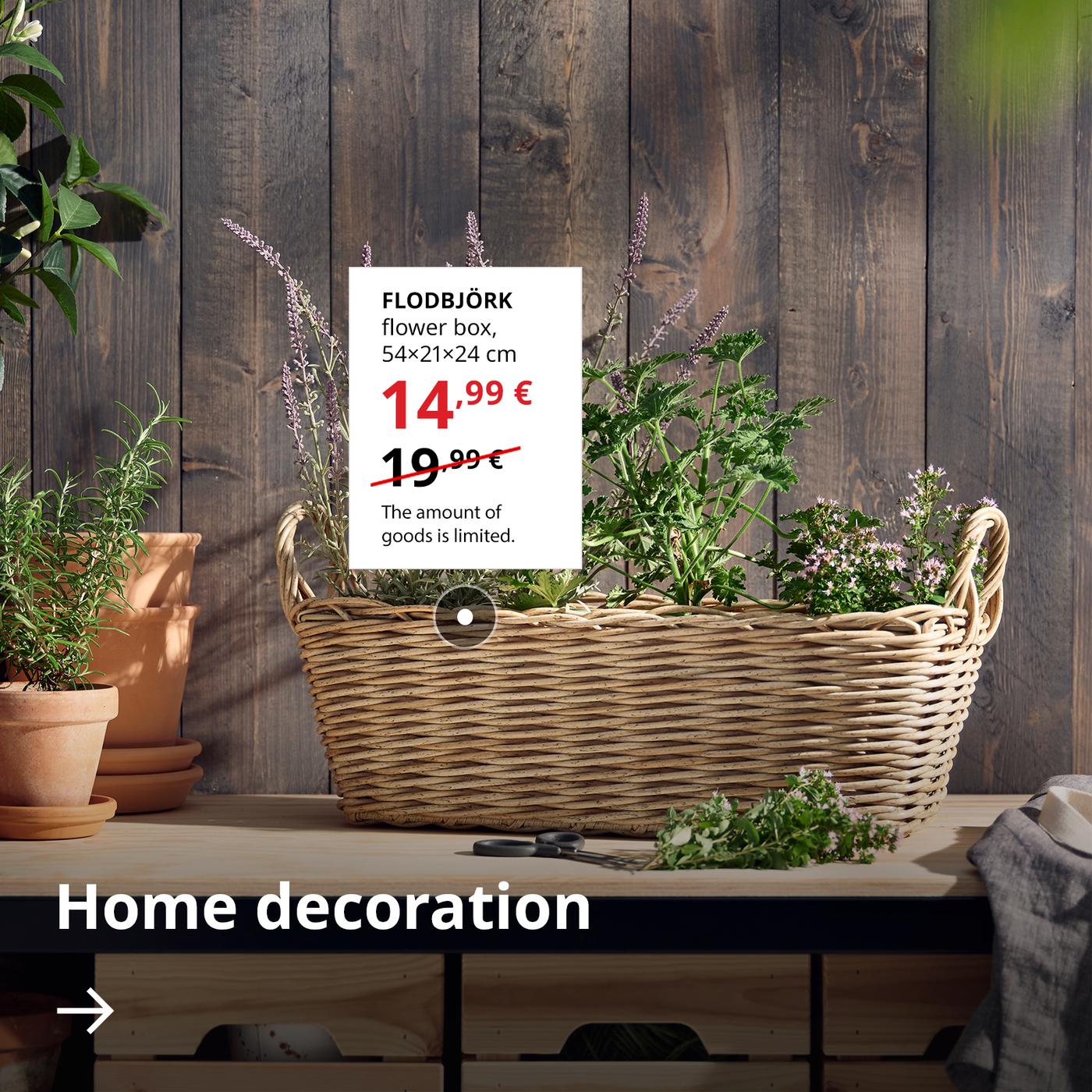 A woven FLODBJÖRK flower box stands by a wooden wall. Next to it, on a white background, the sale price is shown — 14,99 € (regular price 19,99 €). Below, there is an arrow with the text “Home decoration”.