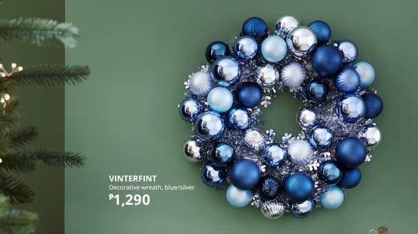 A wreath made from VINTERFINT blue christmas balls of different shades hanging on a green wall beside a lit up Christmas tree.