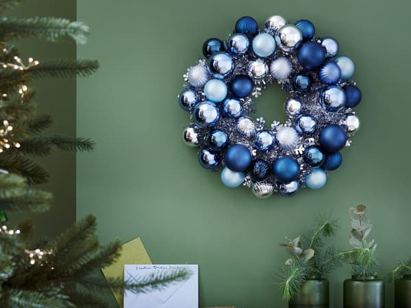 A wreath made from VINTERFINT blue christmas balls of different shades hanging on a green wall beside a lit up Christmas tree.