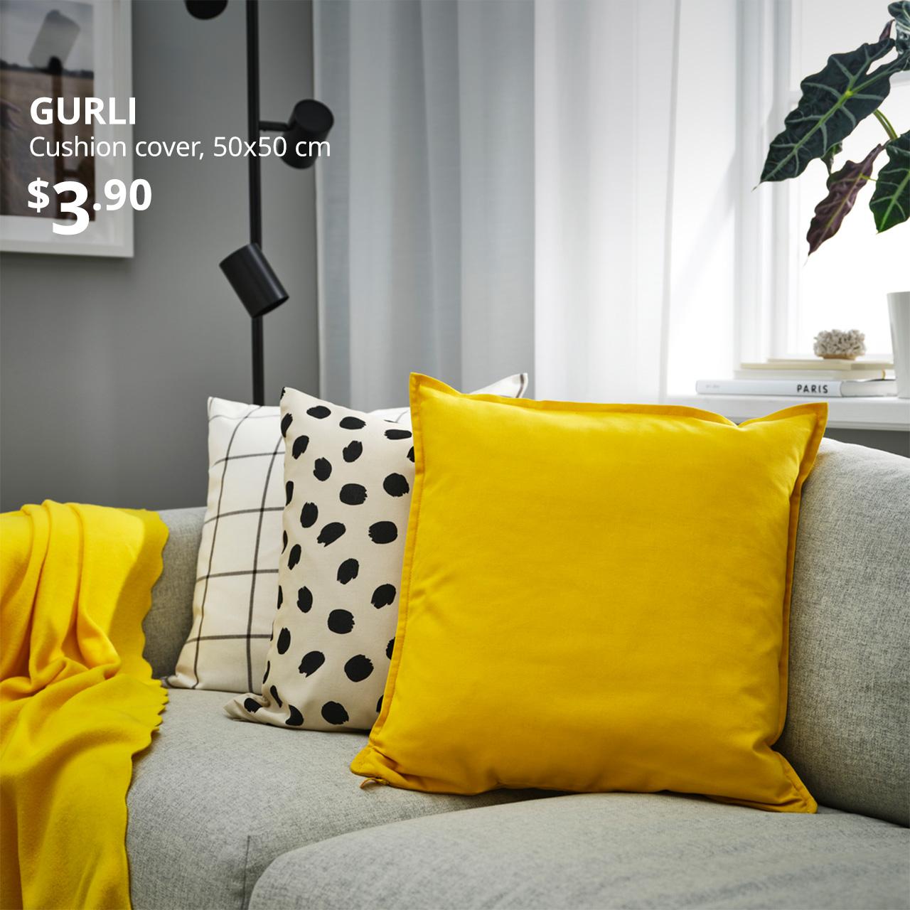 A yellow GURLI cushion cover is used on an inner cushion pad and placed on the faringe light grey FRIHETEN 3-seater sofa bed. White text on top left of the image reads: GURLI, Cushion cover, 50x50 cm. Price: $3.90
