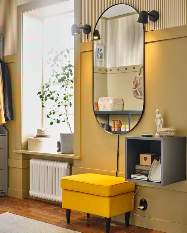 A yellow wall with an oval mirror, two black lamps and a dark grey wall cabinet. A yellow footstool stands under the mirror.
