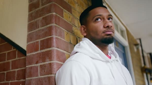 A young bearded man in a white hoodie leaning against a brick wall while looking towards the sky.
