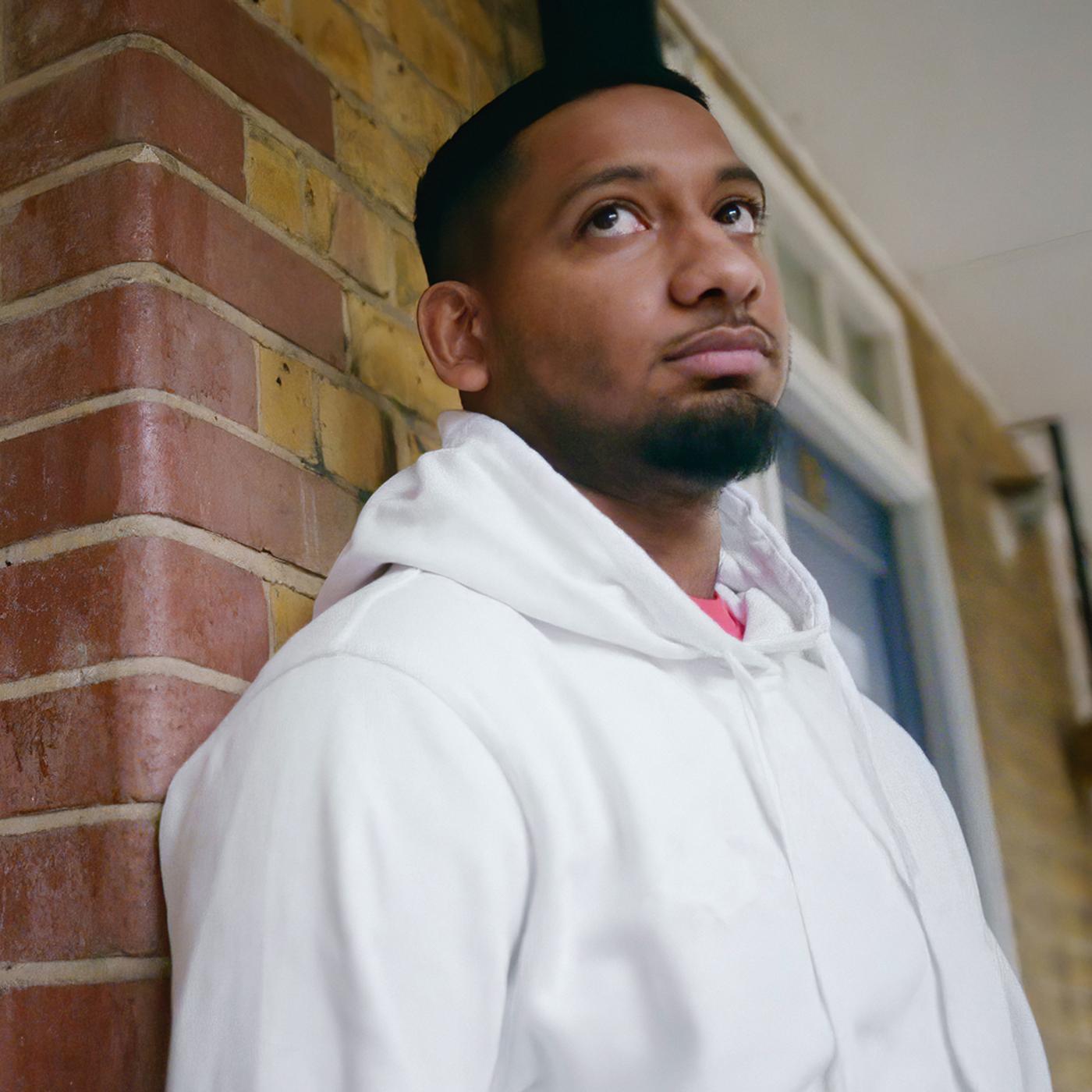 A young bearded man in a white hoodie leaning against a brick wall while looking towards the sky.