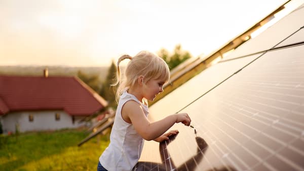 A young blonde girl is touching a solar panel with a straw of grass on the roof of a house in the countryside.