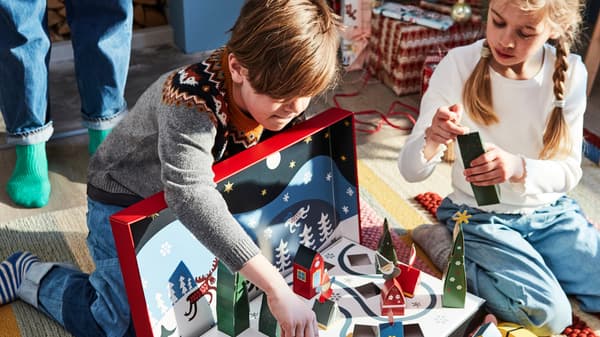 A young boy and girl sit on the floor and assemble a VINTERFINT advent calendar as their mother watches over them.