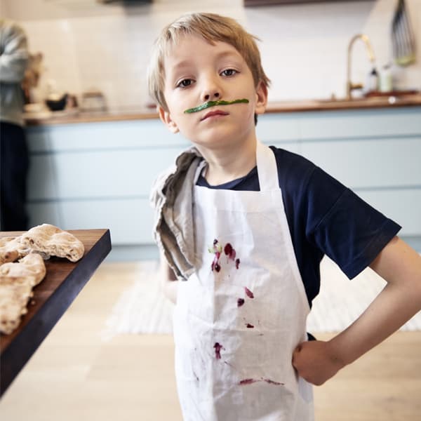 A young boy in the kitchen with an apron covered in food with a play mustache on.