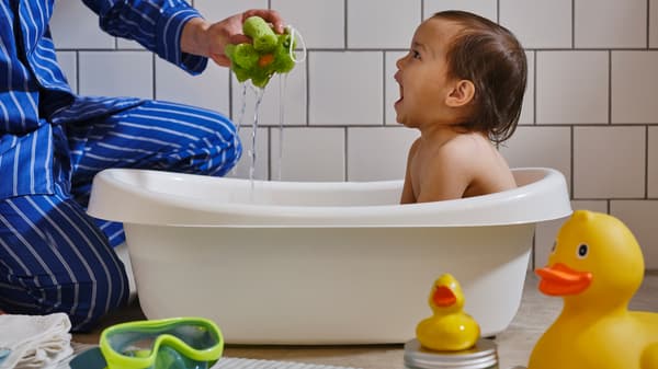 A young child in a white LÄTTSAM baby bath, a white-tiled wall behind, laughs as an adult hand holds up a green sponge.