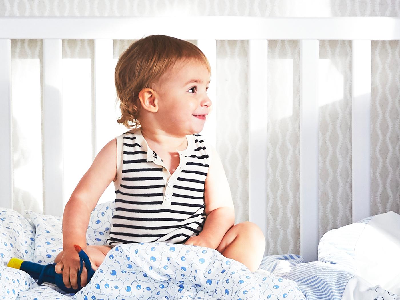 A young child sits in a white SUNDVIK cot with blue and white bed linen with a blueberry pattern, and one side removed.