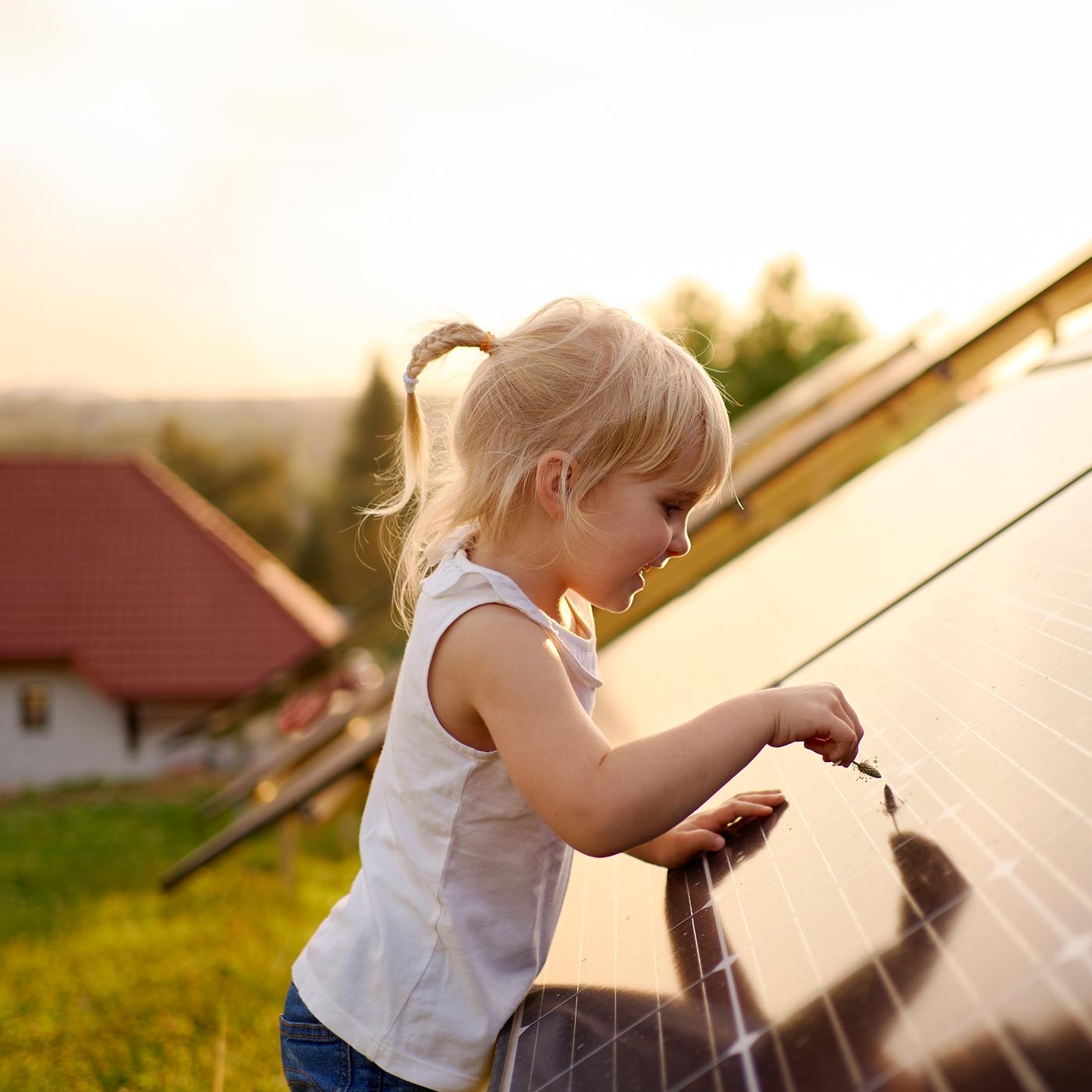A young child with blonde hair runs a grass seed head across the surface of a solar panel while outside in the evening sun.