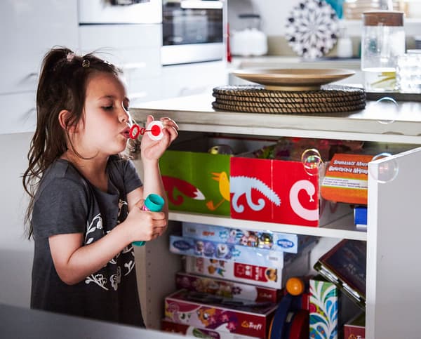 A young girl blowing bubbles in front of an open kitchen unit.